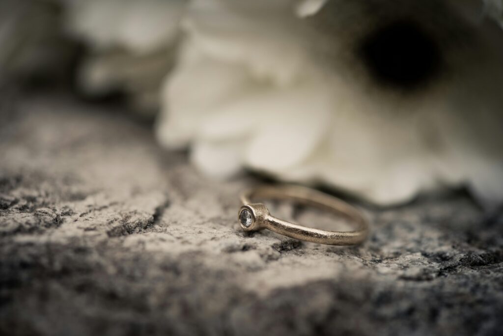 A delicate gold ring with a diamond on a textured stone surface, blurred white flowers in the background.